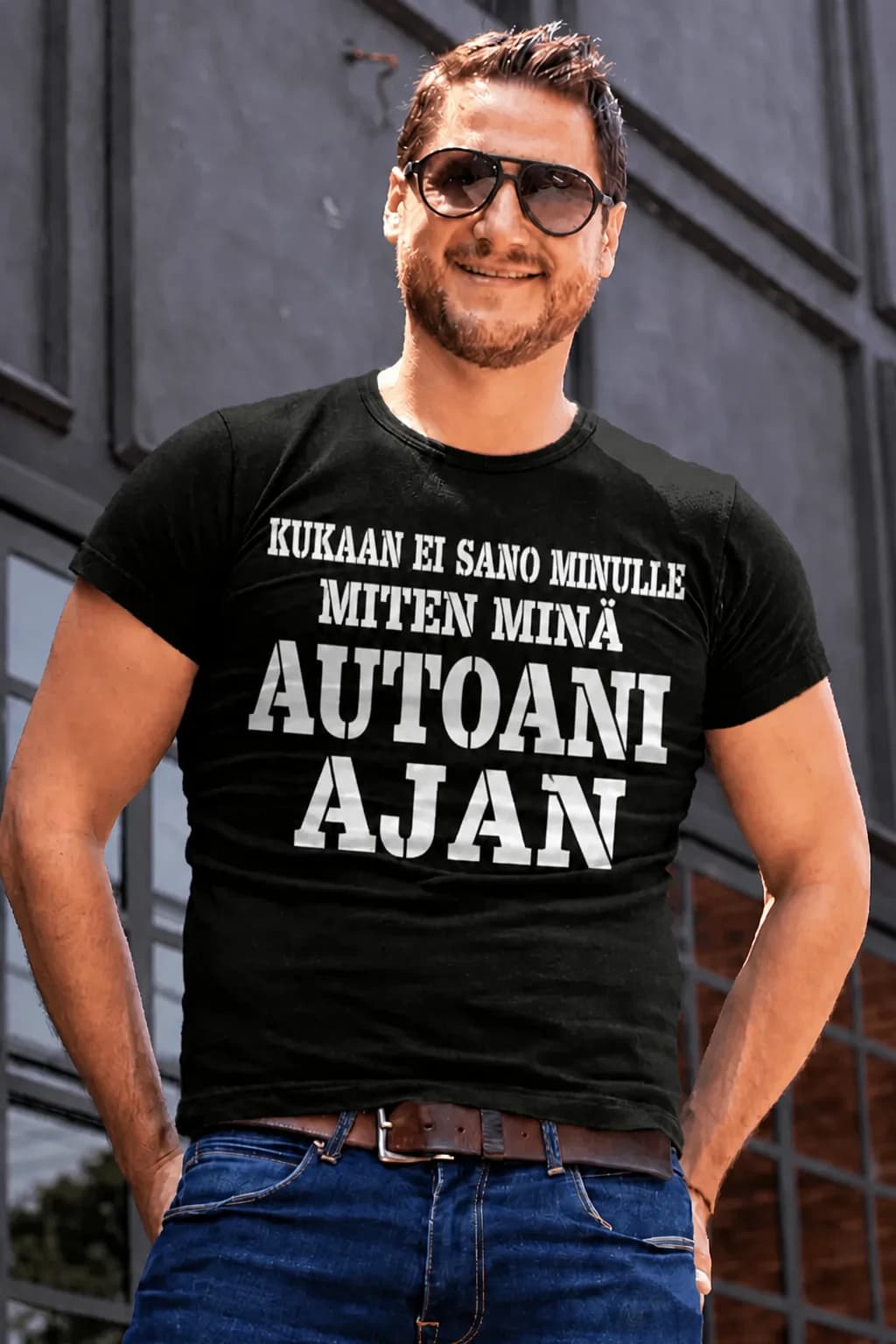 Mies aurinkolaseissa ja mustassa T-paidassa, jossa teksti Kukaan ei sano minulle miten minä autoani ajan