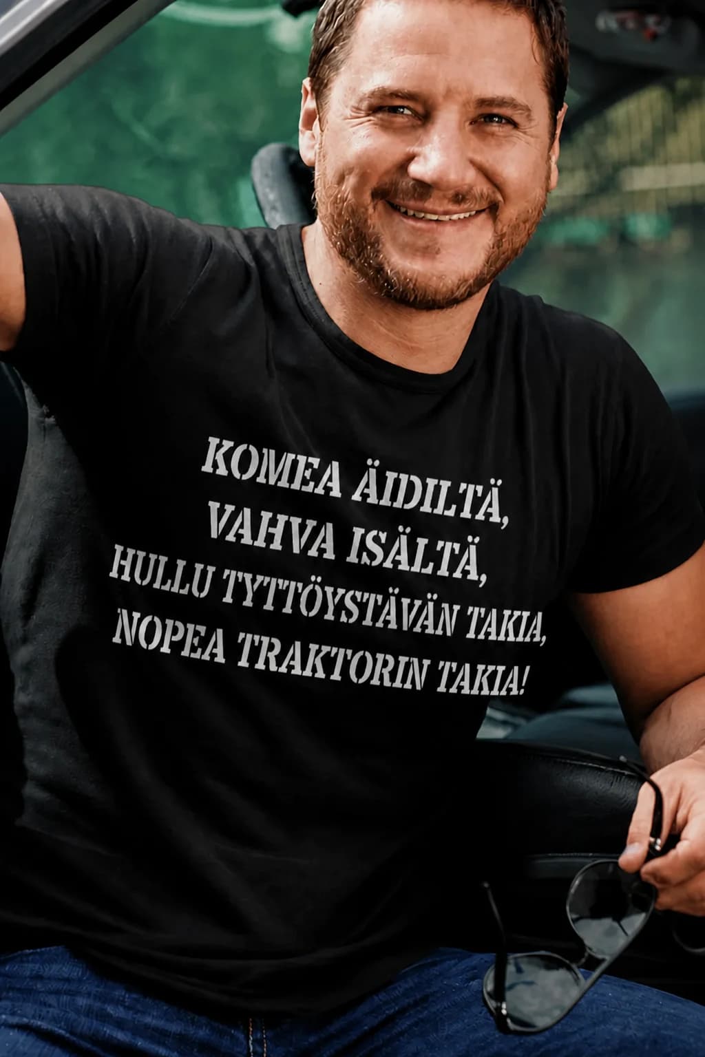 Mies mustassa T-paidassa autoilijalle, jossa lukee komea äidiltä, vahva isältä, hullu tyttöystävän takia, nopea